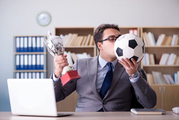 Man in a suit, possibly a Football Agent, kissing a soccer ball while holding a trophy, seated at a desk with a laptop.