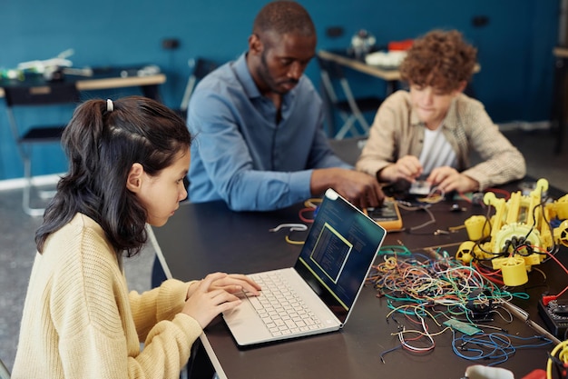 Three people work at a table with a laptop and wiring. No 'Football Agent' elements are present.