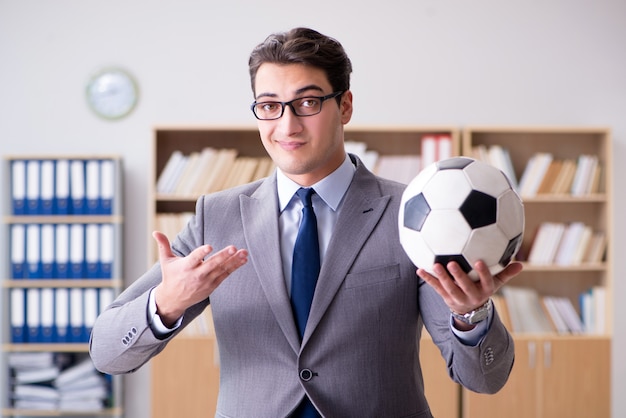 Man in suit holding a soccer ball, gesturing confidently. Bookshelves in the background. Keyword: Football Agent.