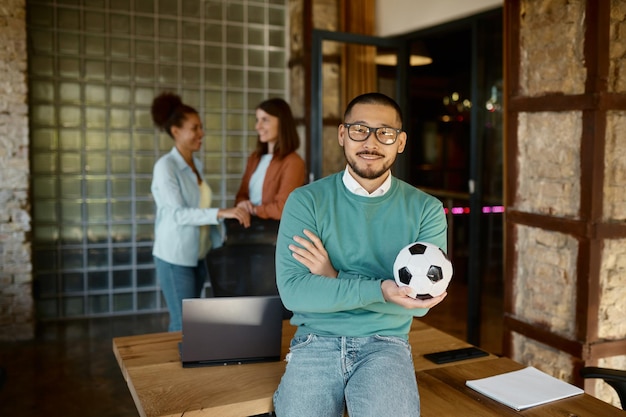 Man holding a soccer ball, representing a Football Agent, leans on a desk. Two women in the background shake hands.