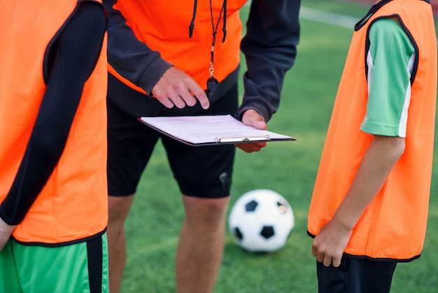 Coach with clipboard instructs two young players in orange vests. Football Agent concept on green field near soccer ball.