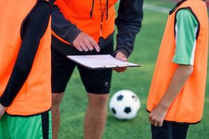 Coach and two young players in orange vests discuss strategy on a clipboard near a soccer ball. Football Agent context.