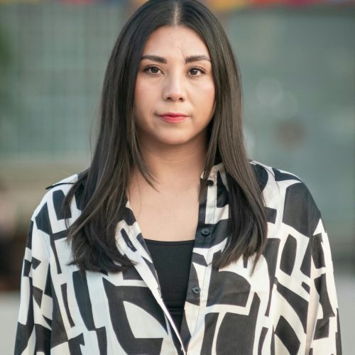 Woman with long hair in a patterned shirt standing in front of a colorful mural. Football Agent.